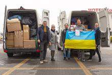 Pięć osób, trzymających ukraińską flagę, stojących przed dwoma busami. Samochodu z tyłu stoją tyłem i mają otwarte drzwi przestrzeni ładunkowej w środku dary.
