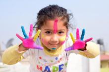 Dziewczynka w wieku przedszkolny ma pomalowane ręce i twarz kolorowymi farbami. 