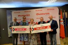 Pięć osób, w tym dwie kobiety, stoją obok siebie i trzymają w rękach dwa duże czeki z kwotą 20 tys. zł i 200 tys. zł. 