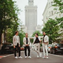 Czterech mężczyzn ubranych w białe garnitury stoi obok siebie na ulicy. W tle widać samochody i wysokie budynki.  
