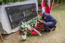  Park. Pomnik ofiar katyńskich. Strażnik miejski składa przed nim wiązankę z białych kwiatów. Obok stoją znicze i trzy biało-czerwone flagi na stojaku. W tle zielone drzewa.