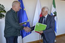 Dwóch mężczyzn przekazuje sobie teczkę na dokumenty. W tle flagi Unii Europejskiej, Polski i Konstancina-Jeziorny.
