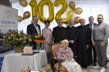 Grupa 7 osób stoi na tle balonów w kształcie liczby 102, na krześle siedzi starsza kobieta.