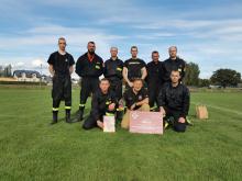 Zdjęcie grupowe 9 strażaków, ubranych w czarne mundury. Stoją w 2 rzędach. Jeden z nich trzyma w rękach duży czek o wartości 1 tys. zł. 