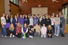 Grupa uczniów w sali konferencyjnej, w tle ściana z czerwonej cegły i ekran do projektora.
