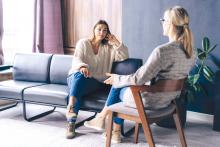 Dwie kobiety siedzą w pomieszczeniu – jedna na krześle, druga na sofie – i rozmawiają.