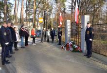 Grupa kilkunastu osób stoi przed pomnikiem pamięci, umieszczonym na ogrodzeniu Białego Dworku. Składają kwiaty i znicze. 