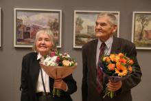 Starsza kobieta i mężczyzna trzymają w dłoniach bukiety kwiatów. Za nimi na ścianie wisza obrazy.