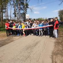 Grupa dzieci na drodze. W drugim planie wysokie drzewa. Przed nimi biało-czerwona wstęga.