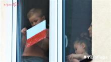 Dziecko stoi w oknie i trzyma w ręku biało-czerwoną flagę. 