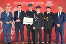 Sześciu mężczyzna, w tym trzech w mundurach strażackich, stoi na tle czerwonej ścianki promocyjnej Państwowej Straży Pożarnej. Jedna osoba trzyma prostokątną tabliczkę, na której widnieje kwota 1 250 000 zł.