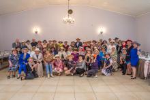 Ponad 60 starszych osób (kobiety i mężczyźni) w dużej sali, ustawionych do zdjęcia. Wszyscy sa w kapeluszach i kolorowo ubrani.