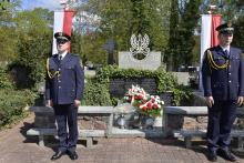 Dwóch mężczyzn w mundurach strazy miejskiej stoi czed grobem, który jest ogrodzeny niskim murkiem. Na tablicy wypisane są imiona i nazwiska oraz  napis „1939, poległym za ojczyznę”. 