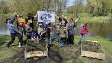 Dziei i dorośli nad brzegiem rzeki. Podnoszą do góry ręce. Z tyły dwóch mężczyzn trzyma plakat z napisem „NASZ BÓBR STOWARZYSZENIE”. Przed nimi donice z ziemia i gałązkami. Jedno dziecko leje na jedną z nich wodę z konewki.     