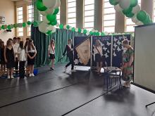Grupa młodzieży na sali gimnastycznej podczas zakończenia roku szkolnego,  poczet sztandarowy, a obok za stolikiem stoi starsza kobieta.Sala jest ozdobiona w zielono-białe balony. Na granatowym przyczepiony jest napis: Coś się kończy coś zaczyna.parawanie