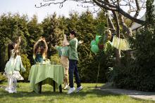 Piatka dzieci stoi wokół stolika z zielonym obrusem. Mają uniesione ręce.  Za nimi rosną drzewa, a obok na gałęzi wiszą nadmuchane balony.