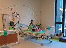 Sala szpitalna. Na ścianie kolorowe rysunki. Na szpitalnym łóżku siedzi dziewczynka. Ma długie włosy i zieloną bluzkę.