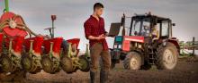 Rolnik trzymający laptopa na tle maszyn rolniczych.