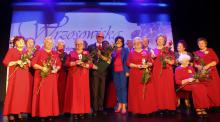Grupa starszych kobiet w czerwonych sukniach stoi na scenie, a wśród nich mężczyzna w garniturze i kobieta w niebieskiej marynarce i spodniach.