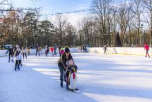 Park zdrojowy, ludzie w strojach zimowych jeżdżą na lodowisku, w tle budynek amfiteatru i drzewa.