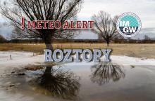 Na górze czerwony napis meteo alert poprzedzony wykrzyknikiem. Obok niego logo Instytutu Meteorologii i Gospodarki Wodnej - niebieski okrąg z białymi literami IMGW, poniżej szary napis roztopy. W tle widać pole z wysokimi drzewami i śnieg leżący na ziemi.s.  