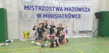 Pięć dziewcząt w czarnych strojach sportowych przyklęka na jednym kolanie i  pozuje do zdjęcia na tle baneru z napisem: mistrzostwa Mazowsza w minisiatkówce.