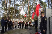 Grupa młodych dziewczyn i chłopców z biało-czerwonymi wieńcami i szarfami oraz trzech starszych mężczyzn i trzy kobiety stoją naprzeciwko tablicy zawieszonej na ogrodzeniu. Przy ogrodzeniu dwóch strażników miejskich w galowych mundurach oraz biało-czerwone flagi.