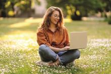 Uśmiechnięta dziewczyna siedzi na łące. Ma długie włosy i jest w okularach. Na kolanach ma otwarty laptop.