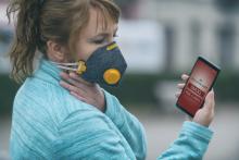 Kobieta z maseczką na twarzy patrzy w monitor telefonu, na który wyświetlony jest napis " Air pollution alert: Smog"