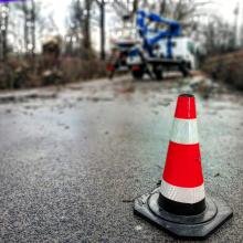 Czerwono-biały pachołek ostrzegawczy na ulicy. W tle widać drzewa i podnośnik do ich ścinania.