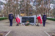 Pośrodku czarna tablica granitowa z tekstem umocowana na prostokątnym kamieniu, po lewej i prawej stronie stoją flagi narodowe, przed tablicą leżą kwiaty i wiązanki oraz palą się znicze