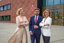 Trzy osoby: dwie kobiety i mężczyzna stoją przed budynkiem z czerwonej cegły
