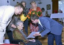 Mężczyzna z listem gratulacyjnym pochylony nad starszą kobietą, w tle inne osoby.