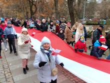 Grupa osób, w tym dzieci i osoby starsze, bierze udział w marszu z okazji Święta Niepodległości. Uczestnicy trzymają dużą, biało-czerwoną flagę Polski, a niektórzy mają przypięte kotyliony.