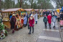 Na zdjęciu widać jarmark bożonarodzeniowy z rzędem kolorowych stoisk i spacerującymi ludźmi w zimowych ubraniach na brukowanej ścieżce wśród drzew.