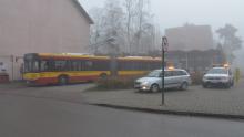 Żółto-czerwony autobus przegubowy warszawskiej komunikacji miejskiej zaparkowany na pętli autobusowej, w otoczeniu budynków i pawilonów handlowych spowitych mgłą. Obok stoją dwa pojazdy techniczne Zarządu Transportu Miejskiego z włączonymi światłami ostrzegawczymi.
