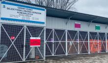 Obiekt zbudowany z elementów metalowych, z frontu widoczne zamykane drzwi z siatką oraz tabliczki z napisami
