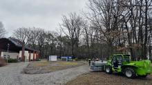 park z zadaszoną sceną, robotników w kamizelkach pracujących przy montażu lodowiska oraz zieloną ładowarkę teleskopową transportującą elementy konstrukcyjne.
