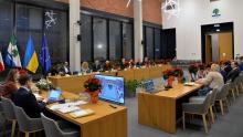 Zdjęcie przedstawia sesję Rady Miejskiej w nowoczesnej sali obrad, z radnymi przy stołach ozdobionych poinsecjami i flagami w tle.