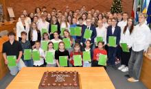 Grupa młodych sportowców trzymających zielone dyplomy pozuje do zdjęcia w sali konferencyjnej z dekoracyjną choinką i tortem z napisem Sportowiec Roku 2024 na pierwszym planie.