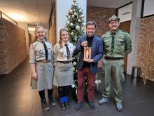 Grupa czterech osób stoi przed udekorowaną choinką, z czego trzy osoby są w mundurach harcerskich, a jedna trzyma zapaloną lampkę.