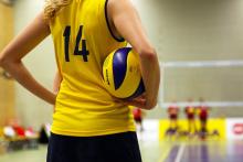Zawodniczka siatkarska w żółtej koszulce z numerem 14 trzyma piłkę do siatkówki, stojąc na boisku przed meczem lub treningiem.