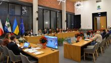 Zdjęcie przedstawia salę obrad z uczestnikami siedzącymi przy stołach, udekorowaną flagami Polski, Ukrainy, Unii Europejskiej oraz roślinami w świątecznym wystroju.