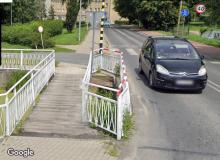 Widok na drewnianą kładkę przez rów wodny, obok samochód.