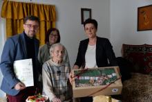 Grupa osób, w tym jubilatka, otrzymuje upominki i gratulacje z okazji setnych urodzin w domowym otoczeniu.