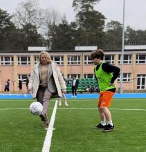 Kobieta w eleganckim stroju kopie piłkę na boisku sportowym, podczas gdy chłopiec w sportowym stroju obserwuje ją z uwagą.