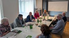 Grupa osób uczestniczy w formalnym spotkaniu przy stole konferencyjnym, omawiając dokumenty i notatki.