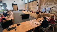 Spotkanie w nowoczesnej sali konferencyjnej, gdzie uczestnicy siedzą przy długich drewnianych stołach, wyposażonych w mikrofony i monitory, uważnie słuchając przemawiających osób.