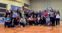 Grupa kobiet w różnym wieku pozuje do wspólnego zdjęcia na sali gimnastycznej, uśmiechając się i wykonując gesty serc lub kciuków uniesionych w górę.