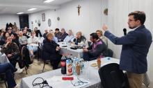 Mężczyzna w marynarce przemawia do zgromadzonych mieszkańców siedzących przy stołach, podczas spotkania w świetlicy wiejskiej.